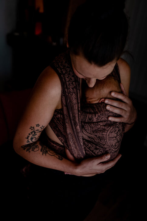 Fotografie einer Mutter. Sie trägt ihr Neugeborenes im Tuch und wiegt es zärtlich in den Schlaf.
