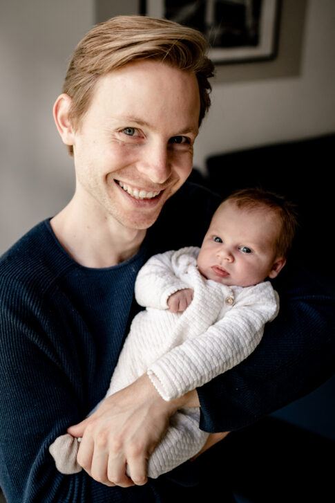 Stolzer Vater lächelt in die Kamera. Er hält sein Neugeborenes im Arm. Dieses Bild entstand beim Familienshooting in Hannover.