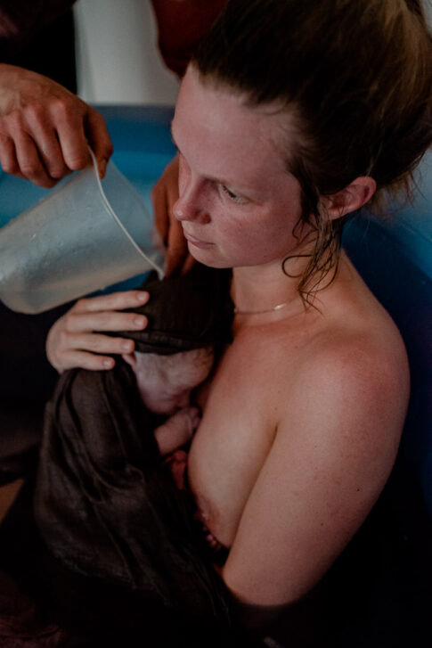 Erschöpfte Mutter nach Geburt im Geburtspool. Doula Wiebke Niemann gießt warmes Wasser über das Neugeborene. Geburtsfotografie Celle Lisa von Rekowski