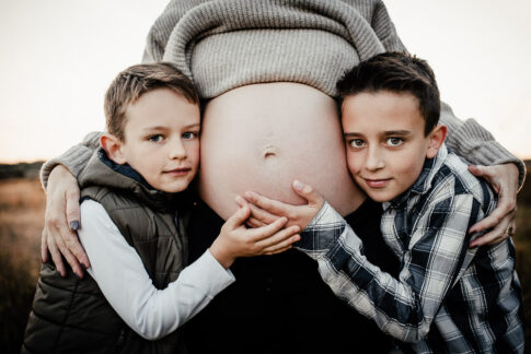 Große Brüder lehnen mit Gesichtern an Babybauch der Mutter. Sie hält die Hände schützend um ihre Söhne.