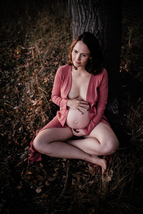 erdverbundene Mutter mit Babybauch lehnt an Baum im Schneidersitz während einer Schwangerschaftsshooting in Wolfsburg
