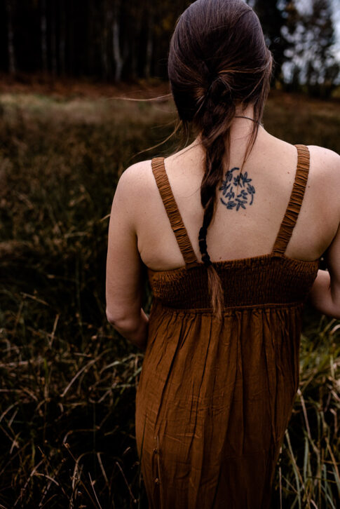 Frau mit Tätowierung auf Rücken im naturfarbenen Kleid geht über Herbstwiese