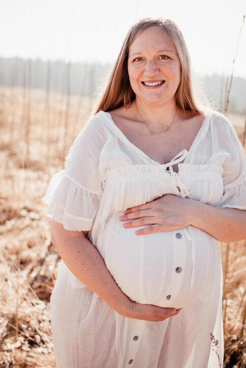 glücklich lächelnde Schwangere in weißem Kleid auf heller Wiese bei Babybauchfotografie in Celle