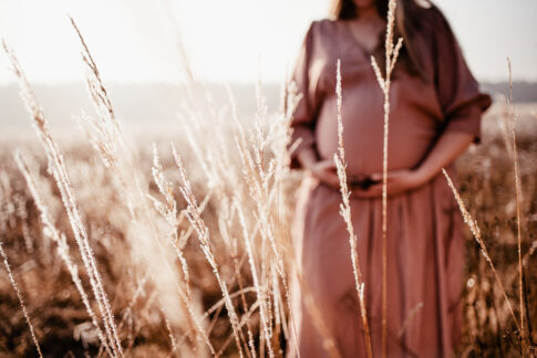Schwangere Frau im Hintergrund unscharf, im Vordergrund Gräser bei Babybauchshooting in Celle